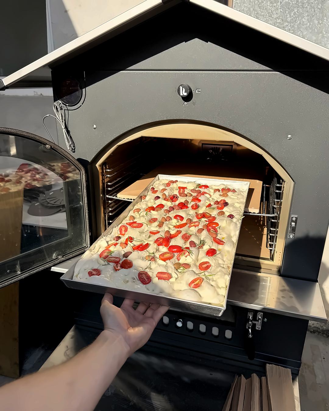 Focaccia mit Kirschtomaten und Rosmarin wird auf dem Blech in den Holzbackofen geschoben